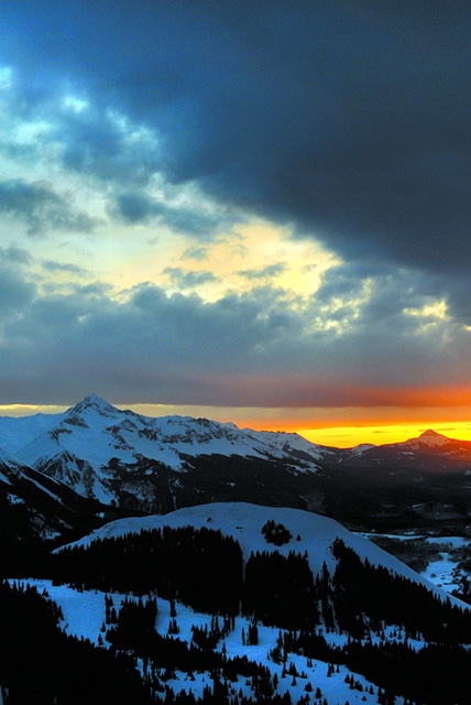 Partly cloudy, dramatic, winter sunset view of Telluride ski area with Mt. Wilson beyond.
