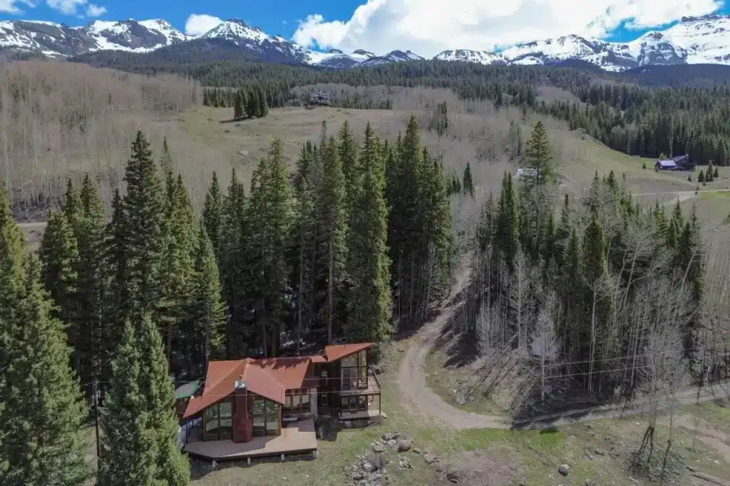 Sunny, aerial view of real estate listing 135 Marmot Way Ophir (Trout Lake), with decks, forest, mountains and blue skies